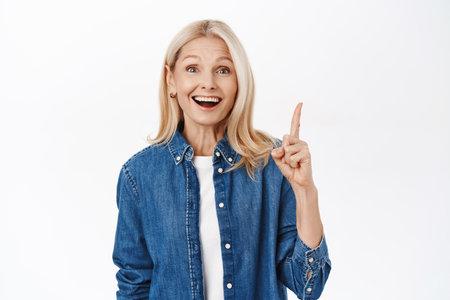 Close up portrait of smiling beautiful lady, senior woman pointing finger up, showing advertisement, top banner, standing over white backgroundの写真素材