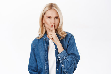 Portrait of blond senior woman shushing, press shh index finger on lips gesture, be quiet, taboo sign, standing over white backgroundの写真素材