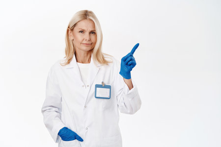 Skeptical woman doctor, smirk displeased, points upper right corner with dissatisfied, doubtful face expression, stands over white backgroundの写真素材