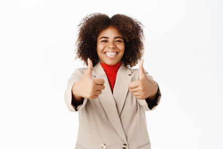 Corporate people. Smiling african american businesswoman, shows thumbs up in approval, praise and compliment, recommends company, stands over white backgroundの写真素材