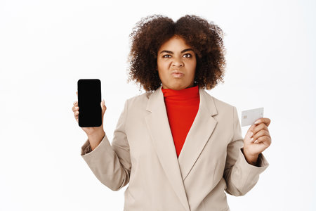 Disappointed african american businesswoman shows telephone screen, mobile interface and credit card, express dislike, stands over white backgroundの写真素材