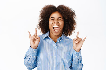 Carefree african american woman, shows rock on, heavy metal gesture and smiling, express positivity and happiness, standing over white backgroundの写真素材