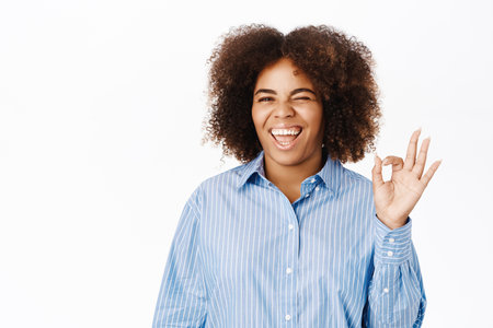 Portrait of smiling african american woman shows okay, ok sign, approve excellent choice, recommends smth, stands over white backgroundの写真素材