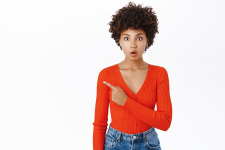 Smiling african american woman in stylish outfit, pointing finger left, showing promotion text on copy space, empty banner, stands over white backgroundの写真素材
