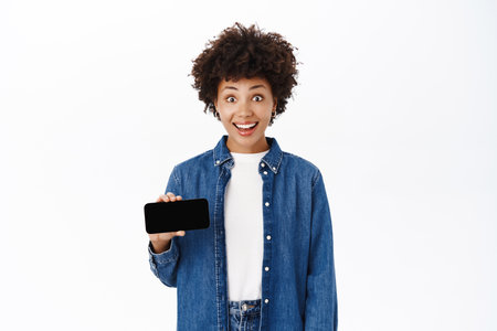 Happy young woman smiles, shows her mobile phone screen, demonstrates website or online shop on her smartphone, white backgroundの写真素材