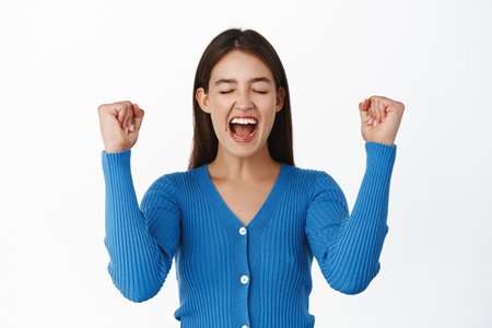 Image of enthusiastic young woman winning, shouting with joy and fist pump, achieve goal success, celebrating victory, standing over white backgroundの写真素材