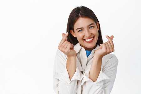 Cute smiling girl showing hand hearts and looking romantic at camera, standing over white backgroundの写真素材