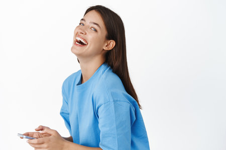 Portrait of happy modern girl with phone, smiling and laughing, looking carefree, wearing blue t-shirt. Celullar company advertisementの写真素材