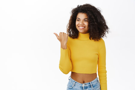 Enthusiastic young african woman pointing left, cute girl student showing advertisement, logo or banner beside her, smiling amazed, standing over white backgroundの写真素材