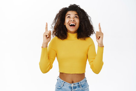 Enthusiastic young african woman, pointing finger up, showing advertisement, top promo banner, logo upwards, standing over white backgroundの写真素材