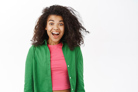 Portrait of black curly girl looks with happy face at camera, surprised by smth, amazed by big news promo, white backgroundの写真素材