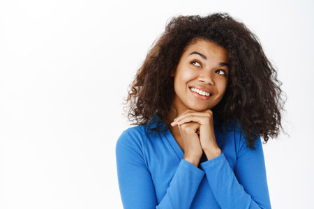 Portrait of beautiful young Black woman smiling, looking happy and carefree, daydreaming, standing in casual clothes over white backgroundの写真素材