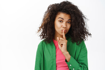 Shh keep it secret. Young teenage curly girl, makes hush, shush taboo sign, press one finger to lips and looks mysterious, white backgroundの写真素材