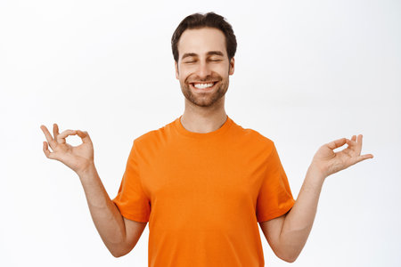 Meditation and mindfulness. Man meditating, practice yoga, reaching nirvana, feeling relaxed, standing in orange tshirt over white backgroundの写真素材