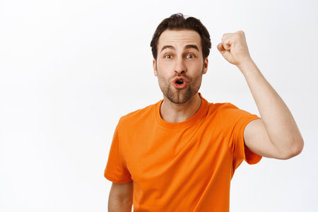 Enthusiastic man raising fist up in support, cheering and chanting, watching sports game, standing in orange tshirt over white backgroundの写真素材
