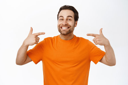 Portrait of handsome man with perfect smile, pointing at his teeth, standing in orange tshirt, white studio backgroundの写真素材