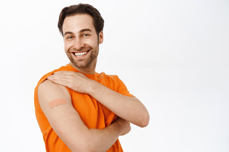 Covid vaccination campaign. Smiling healthy adult man shows his shoulder  from coronavirus vaccine shot, standing in orange tshirt over white backgroundの写真素材