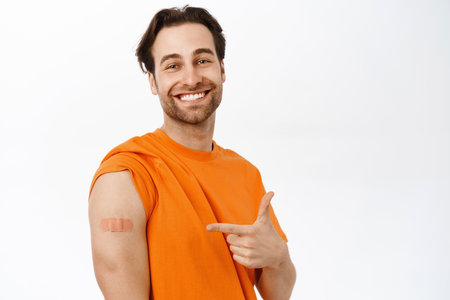 Covid vaccination campaign. Smiling healthy adult man shows his shoulder from coronavirus vaccine shot, standing in orange tshirt over white backgroundの写真素材