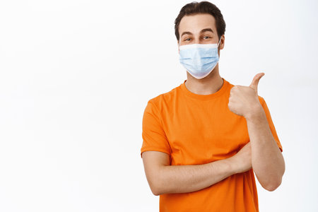 Happy smiling man wears medical face mask, shows thumbs up, recommends using preventive measures, protection from covid 19, white backgroundの写真素材