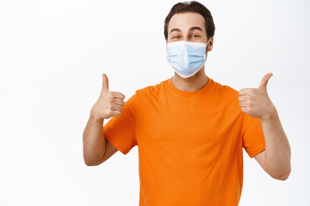 Happy smiling man wears medical face mask, shows thumbs up, recommends using preventive measures, protection from covid 19, white backgroundの写真素材