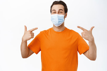Image of smiling man points at his face with medical face mask, wears orange t-shirt, white backgroundの写真素材