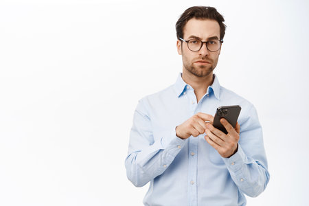 Portrait of handsome businessman using mobile phone, reading on smartphone, working on calls, standing over white backgroundの写真素材