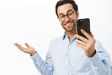 Handsome businessman demonstrating smth on video call. Office worker talking on mobile phone, showing smth behind him, standing over white backgroundの写真素材