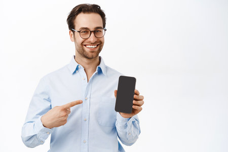 Handsome smiling brunette man pointing at his mobile phone screen. Entrepreneur showing smth on smartphone, recommending application, white backgroundの写真素材