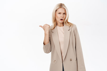 Shocked mature woman, 50 years old, middle aged lady pointing finger left, looking offended and concerned, standing over white backgroundの写真素材