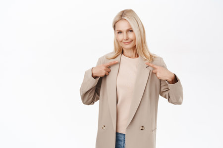 Smiling blond middle aged woman, mature lady in her 50s, pointing fingers at herself, wearing trench coat, standing over white backgroundの写真素材