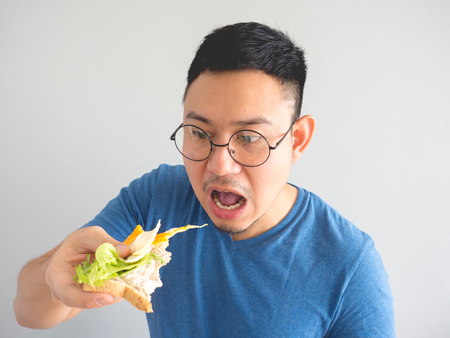 Asian man eat sandwich with vegetable and egg and make funny face.の写真素材