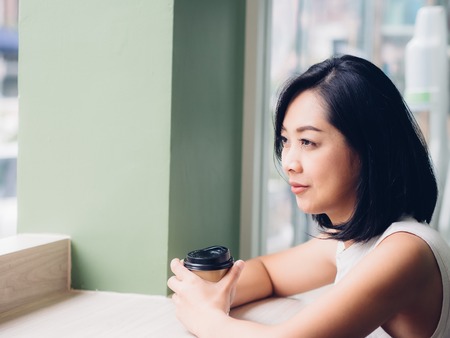 Asian woman is drinking a hot cup of coffee and sit by the windows.