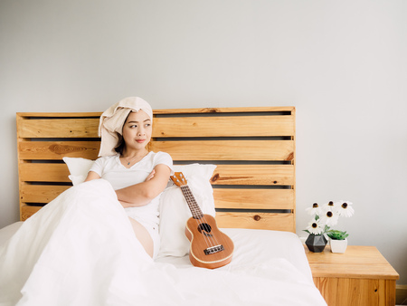 Asian woman has a fine relax day with her ukulele on bed.の写真素材