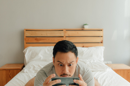 Asian man is playing mobile game with his smartphone on the bed.の写真素材