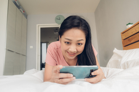 Happy Asian woman is playing mobile game on her bed.