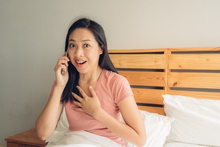 Shocked and surprised Asian woman is having a phone conversation on her bed.の写真素材