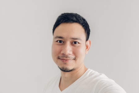 Smile Asian man is taking selfie of himself with white t-shirt and grey background.の写真素材