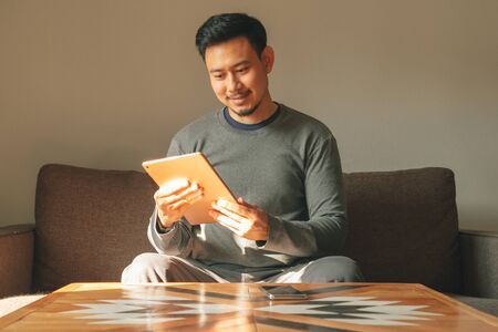Asian man is using his smart tablet device in his living room apartment.