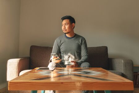 Asian man sits on the sofa in his living room, feeling relax in the morning.