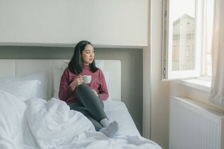 Asian woman is drinking hot morning coffee on her bed int the apartment.の写真素材
