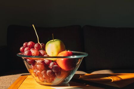 Glass bowl of variety of fruits on wooden table in warm sun light.