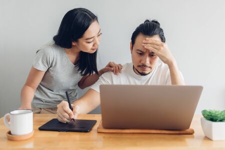 Serious Asian man is working online with his wife support in concept of work from home.