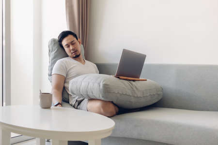Sleepy Asian man working on his laptop on the sofa in his apartment.の写真素材