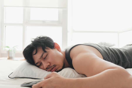 Sleepy Asian man is using smartphone while lying on the bed.の写真素材
