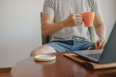 Man is picking up an orange coffee cup while working on his laptop.
