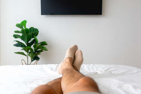 View of mans legs on white bed with TV and plant. Concept of relaxation.の写真素材