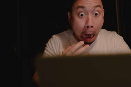 Shocked and surprised face of Asian man using laptop alone at night.