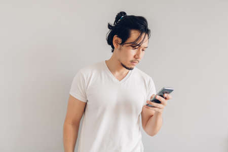 Serious Asian man in white t-shirt is using smartphone on grey background.の写真素材
