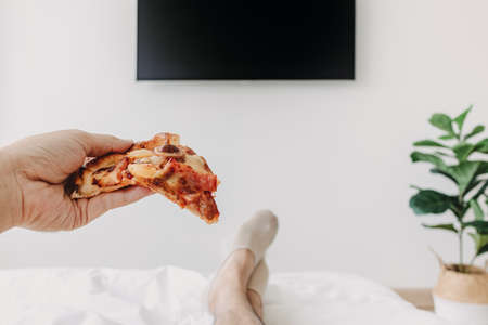 Person on bed and eating pizza watch television. Concept of stay home.