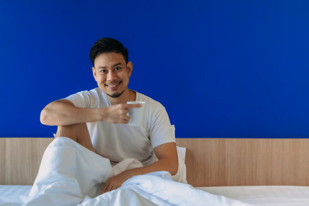 Asian man drinks morning coffee on the bed with a blue screen background.の写真素材